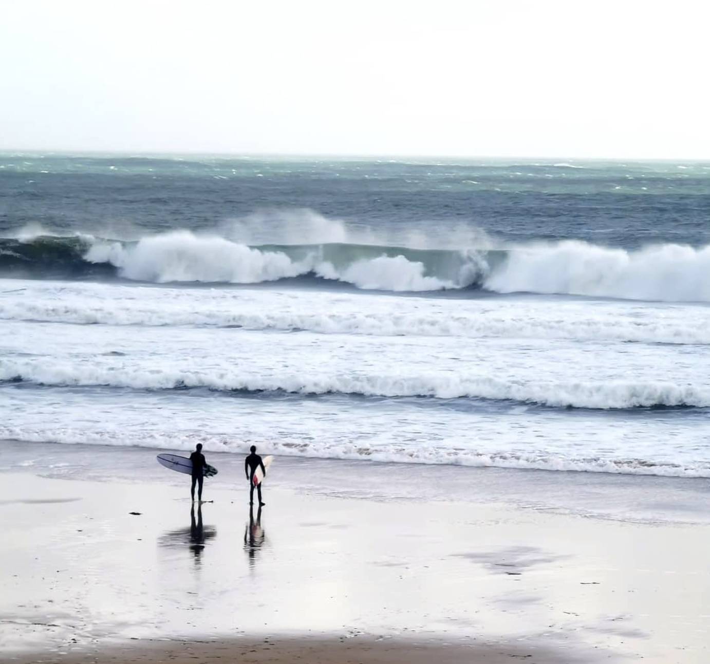 Two surfers in wetsuits on a wet beach, looking out at breaking waves